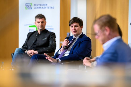 Staatsminister Conrad Clemens sitzt neben Tomi Neckov und Maximilian Schulyok auf einem Podium auf der Bühne und spricht in ein Mikrofon