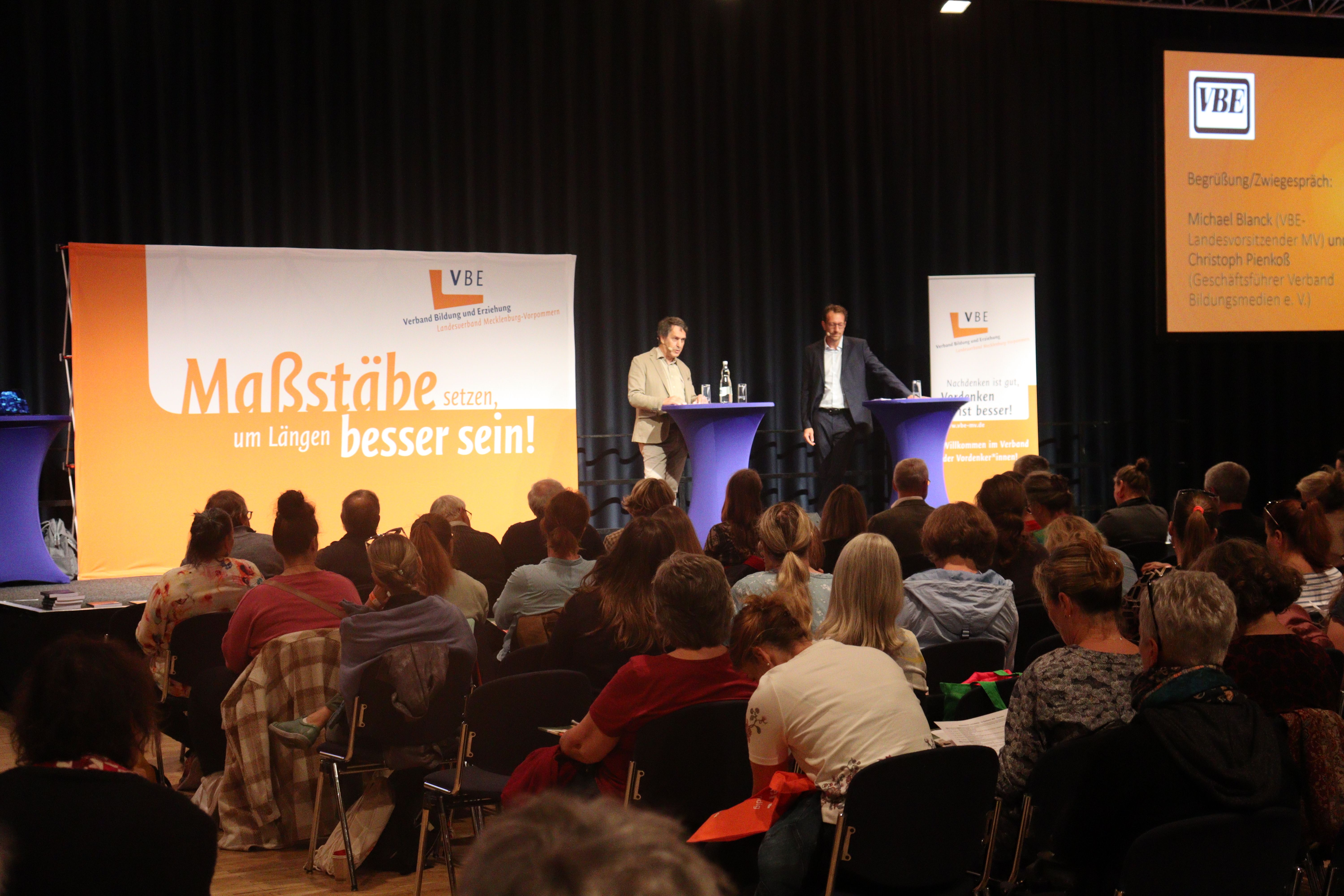 Michael Blanck und Christoph Pienkoß stehen auf einer Bühne vor Zuhörerinnen und Zuhörern.