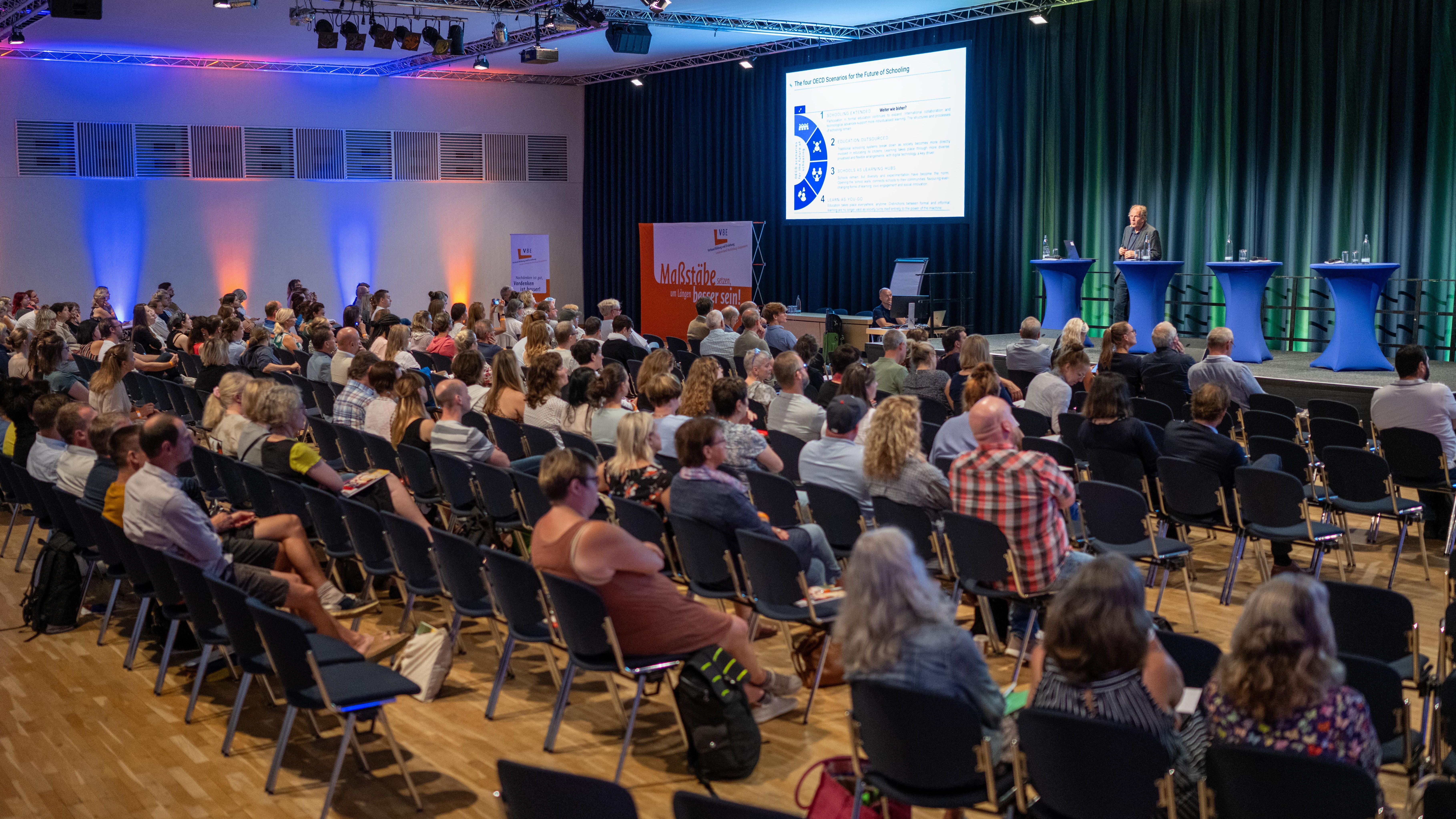 Ein Referent spricht von der Bühne herunter zu voll besetztem Publikumsraum